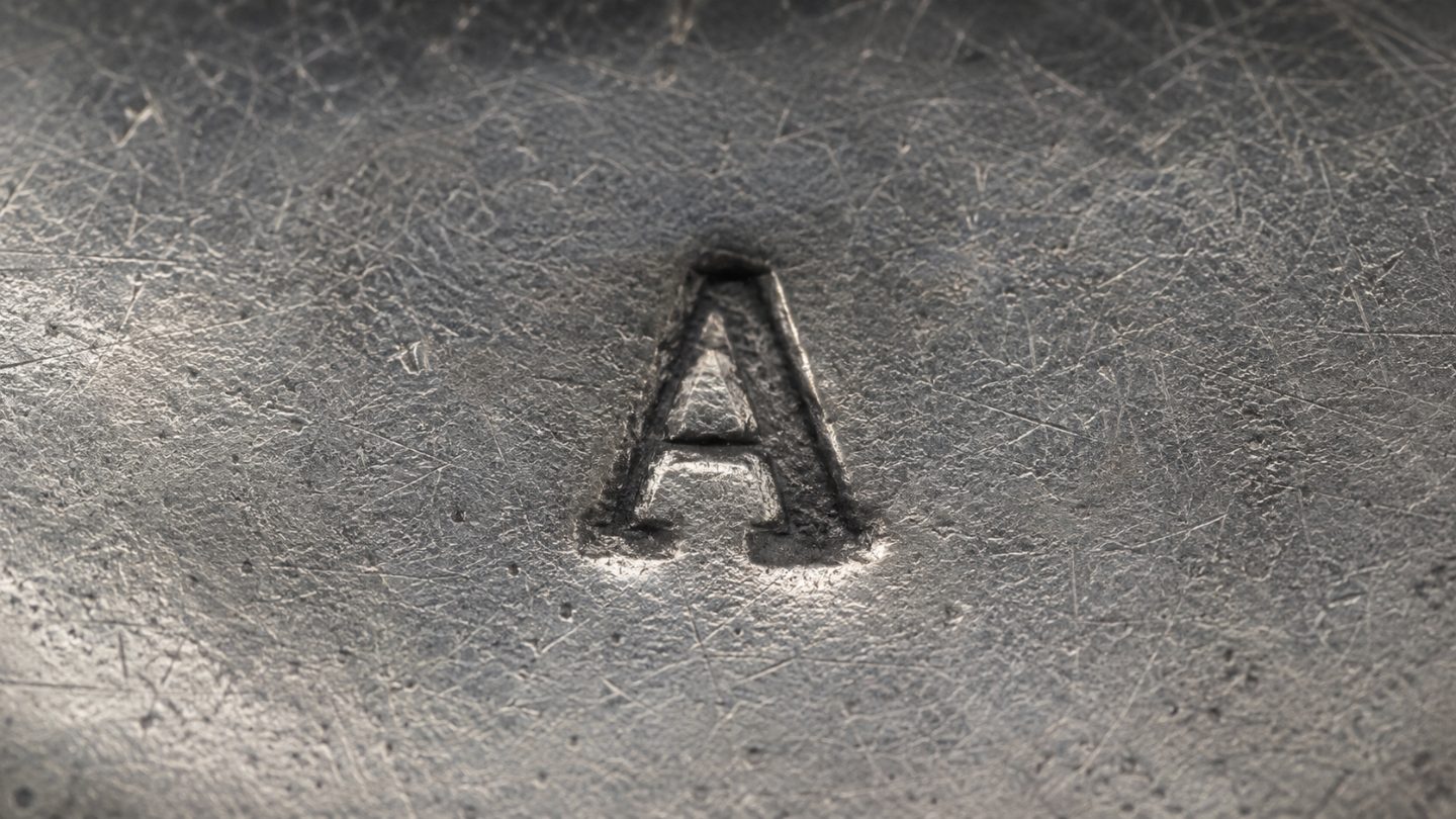 A maker's mark struck into silver: the letter A impressed into the metal, surrounded by fine tool marks and scratches.